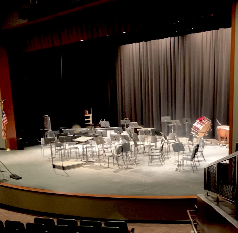 An elegant band stage set in a large theater, featuring rows of chairs and music stands against black curtains with wood trim. The stage is softly lit, evoking a sense of ambiance and anticipation for a performance of "Arabian Nights"